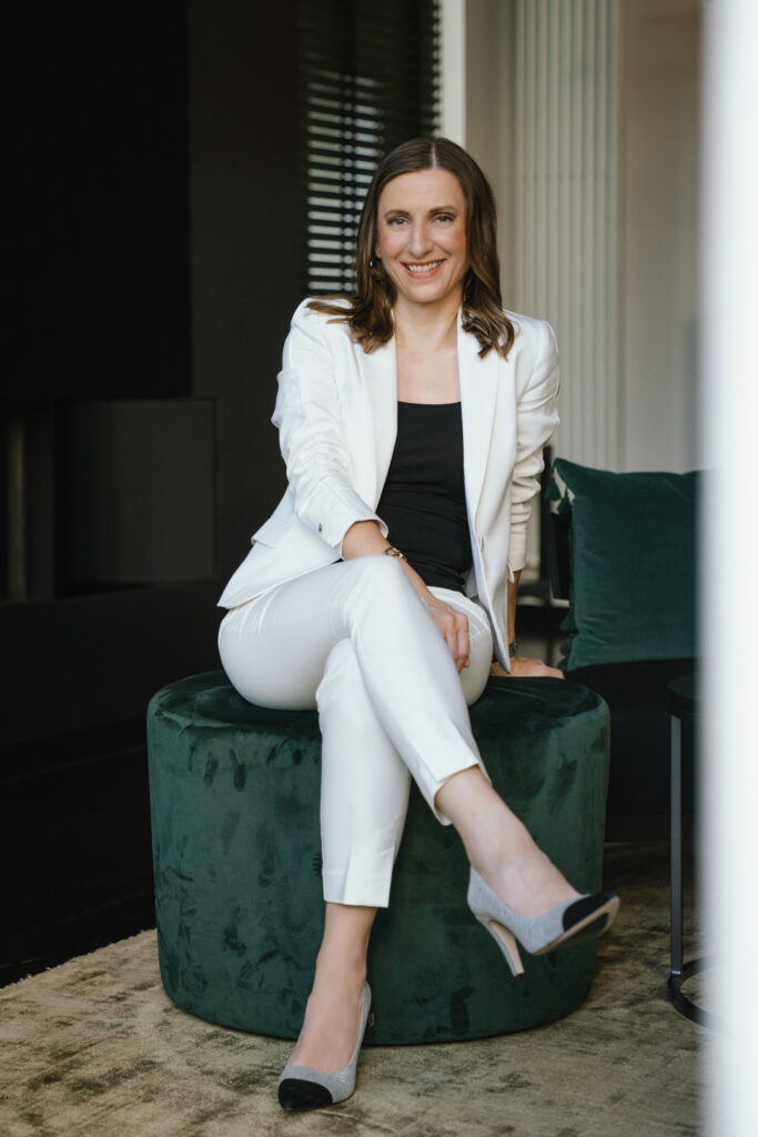 Eine Frau im weißen Anzug sitzt auf einer grünen Ottomane und lächelt mit gekreuzten Beinen in einer modernen Inneneinrichtung - die perfekte Inspiration für einen Hochzeitsplaner, der elegante Hochzeiten an der Ostsee plant.