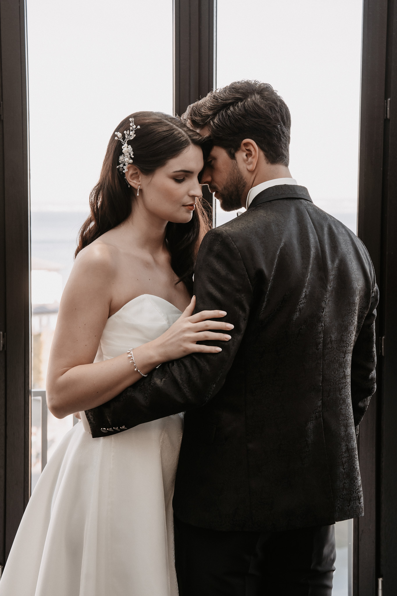 Braut und Bräutigam umarmen sich zärtlich an einem Fenster und berühren sich in einem intimen Moment, während das weiche Licht an eine romantische Flucht nach Rügen an der Ostsee erinnert.
