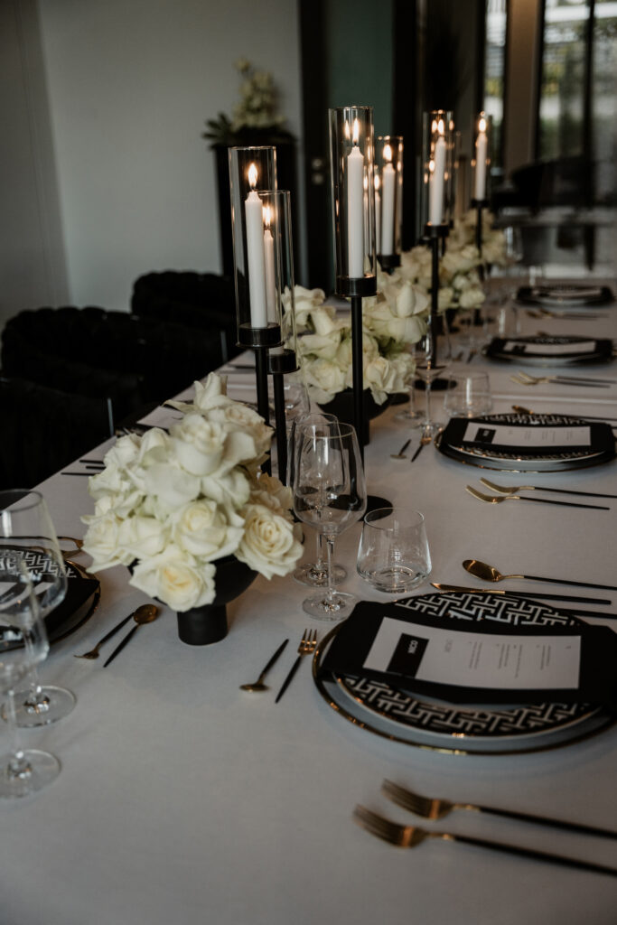 Elegant gedeckter Tisch mit weißen Rosen, schwarzen Kerzenleuchtern und schwarz-goldenen Tellern auf einer weißen Tischdecke - die perfekte Inspiration für einen Hochzeitsplaner auf Sylt oder Rügen.