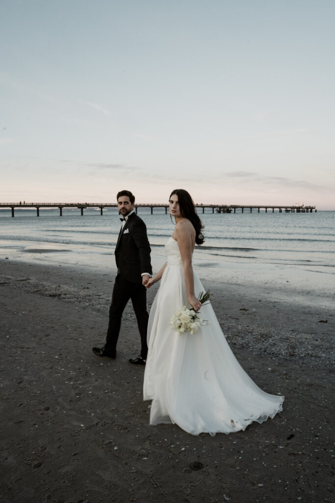 Braut und Bräutigam halten sich an den Händen und spazieren am Strand bei Sonnenuntergang, mit einer Seebrücke im Hintergrund - perfekt für eine romantische Ostsee-Hochzeit, geplant von einem erfahrenen Hochzeitsplaner.