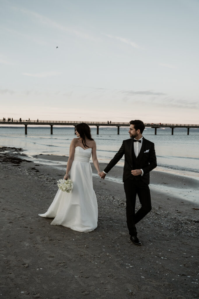 Braut und Bräutigam gehen Hand in Hand an einem Sylter Strand in der Nähe des Wassers bei Sonnenuntergang, mit einer Seebrücke im Hintergrund.