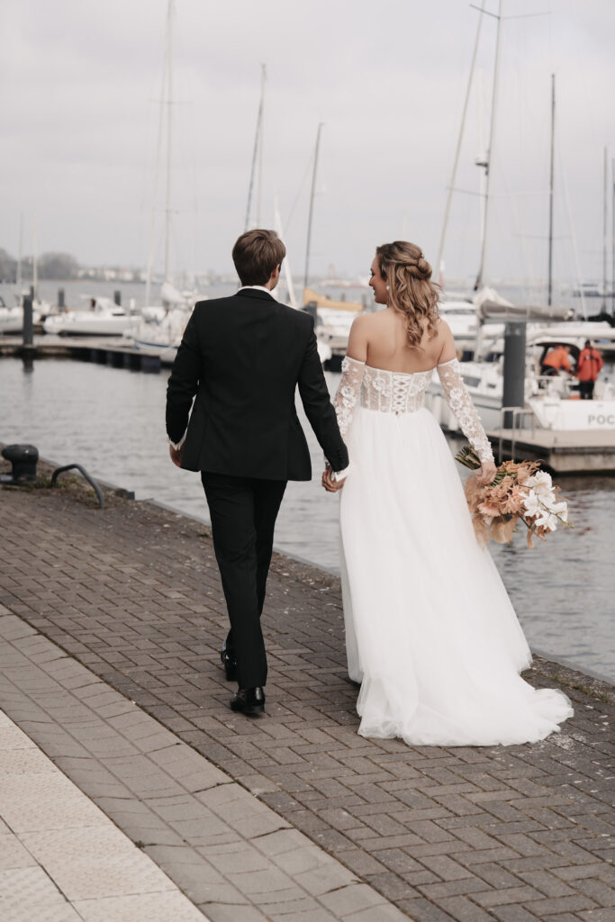 Eine Braut und ein Bräutigam gehen Hand in Hand durch einen Yachthafen mit Segelbooten auf Sylt, von der Kamera abgewandt.