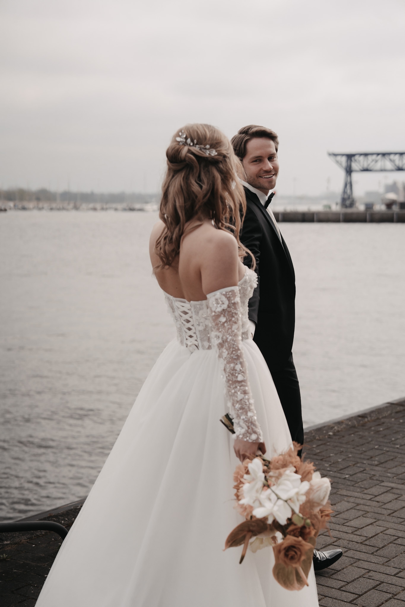 Braut im Spitzenhochzeitskleid hält Blumen und geht mit einem lächelnden Bräutigam im Anzug an der Ostsee spazieren.