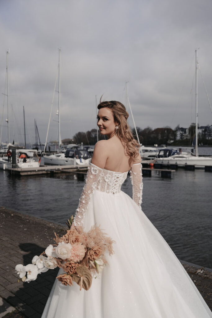 Die Braut in einem schulterfreien Kleid mit einem Blumenstrauß in der Hand steht an einem Yachthafen auf Rügen, mit Booten und Wasser im Hintergrund - eine idyllische Szene für jeden Hochzeitsplaner.