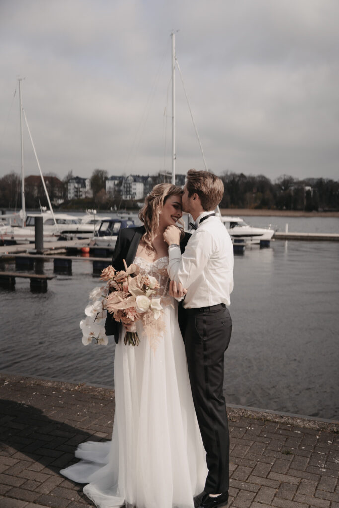 Braut und Bräutigam umarmen sich an einem Yachthafen auf Sylt, mit Booten und der Ostsee im Hintergrund.