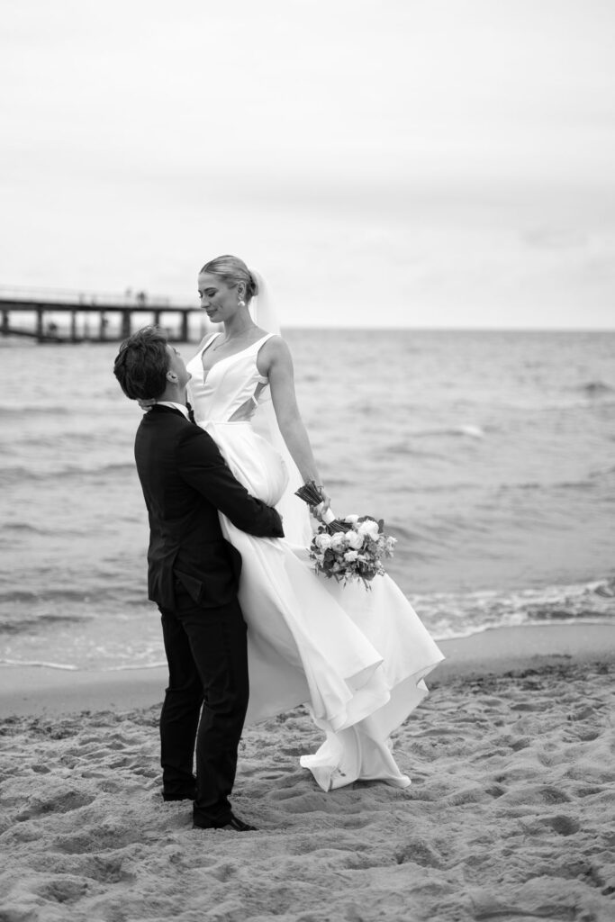 Ein Bräutigam hebt seine Braut lächelnd auf den Sandstrand der Ostsee, mit dem Meer und der Seebrücke im Hintergrund - ein unvergesslicher Moment, eingefangen vom Hochzeitsplaner auf Sylt.