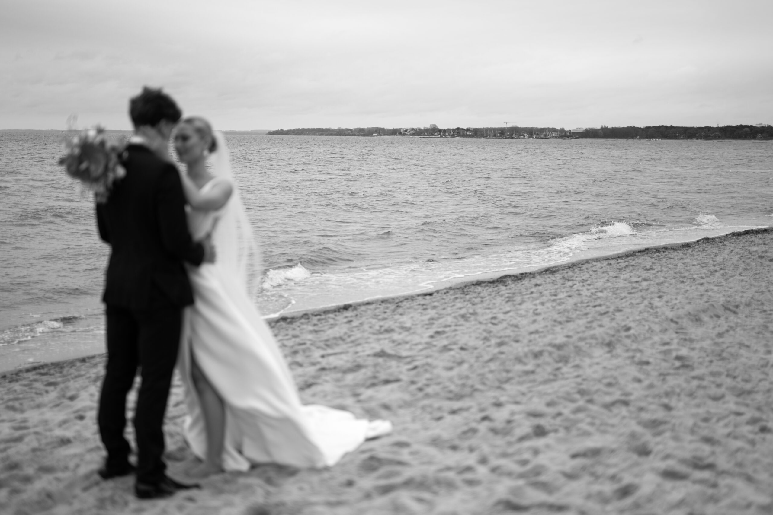 Eine Braut und ein Bräutigam stehen an einem Sandstrand mit der Ostsee im Hintergrund und umarmen sich gegenseitig.