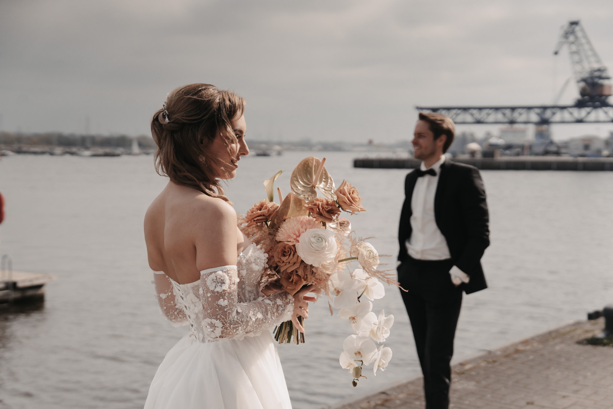 Eine Braut mit einem Blumenstrauß in der Hand steht am Wasser auf Rügen, im Hintergrund ein Bräutigam im Smoking, der die Romantik an der Ostsee einfängt.