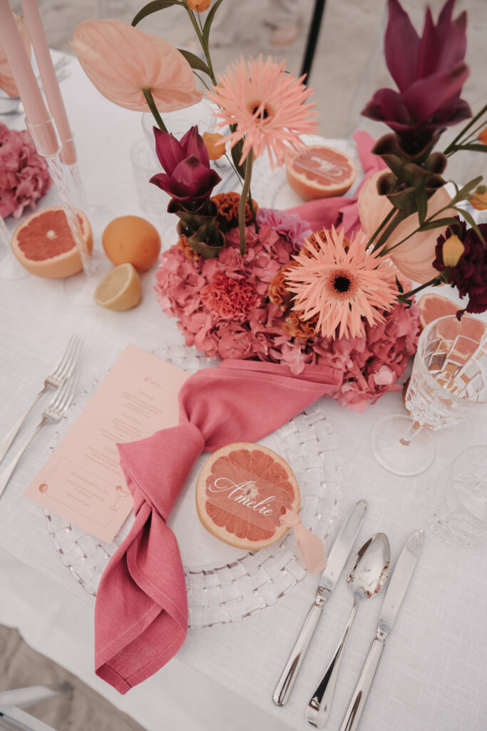 Elegant gedeckter Tisch mit rosafarbenen Blumen, Zitrusfrüchten und einer Grapefruit-Tischkarte auf einer weißen Tischdecke - perfekt für ein sommerliches Fest, inspiriert vom Charme Sylts und der erfrischenden Atmosphäre der Ostsee.