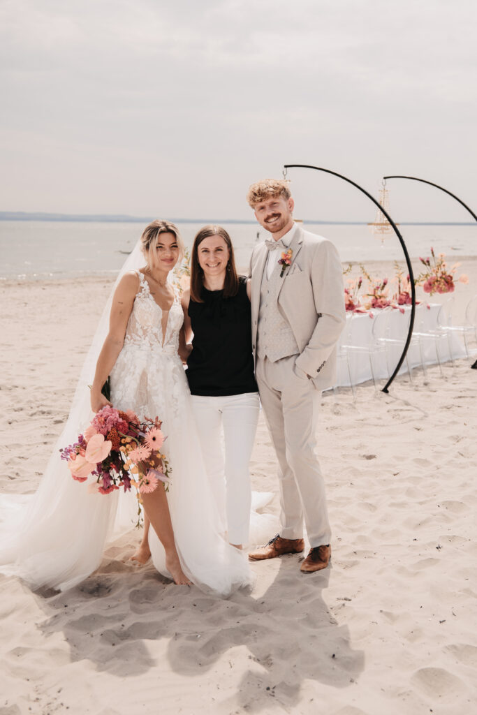 Braut, Bräutigam und eine Frau - möglicherweise ihre Hochzeitsplaner - posieren zusammen an einem Sandstrand auf Sylt mit einer blumigen Hochzeitsdekoration im Hintergrund.