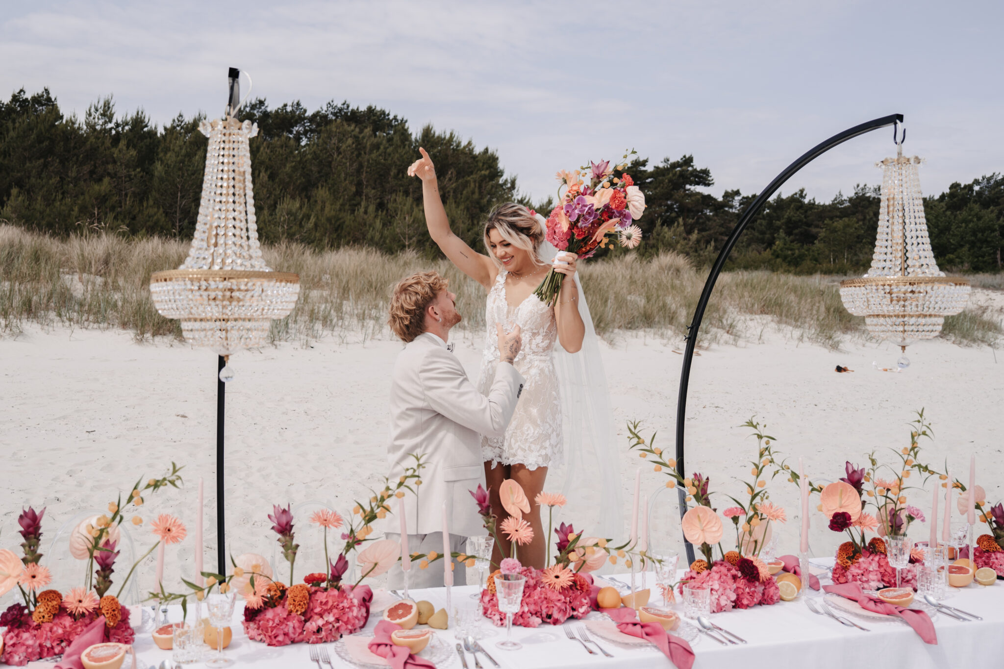 Ein Mann macht einer Frau an einem mit Blumen und Kronleuchtern geschmückten Strandtisch einen Heiratsantrag, während ein Hochzeitsplaner jedes Detail für diesen unvergesslichen Moment an der Küste von Sylt arrangiert.