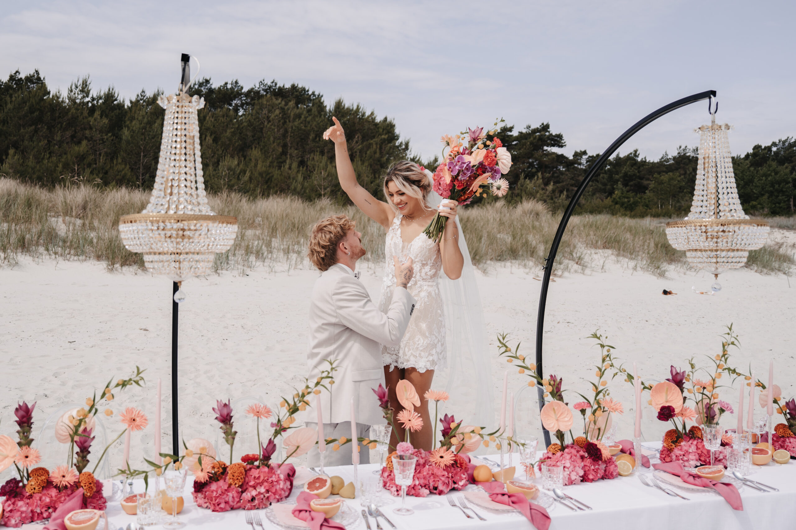 Ein Mann macht einer Frau an einem mit Blumen und Kronleuchtern geschmückten Strandtisch einen Heiratsantrag, während ein Hochzeitsplaner jedes Detail für diesen unvergesslichen Moment an der Küste von Sylt arrangiert.