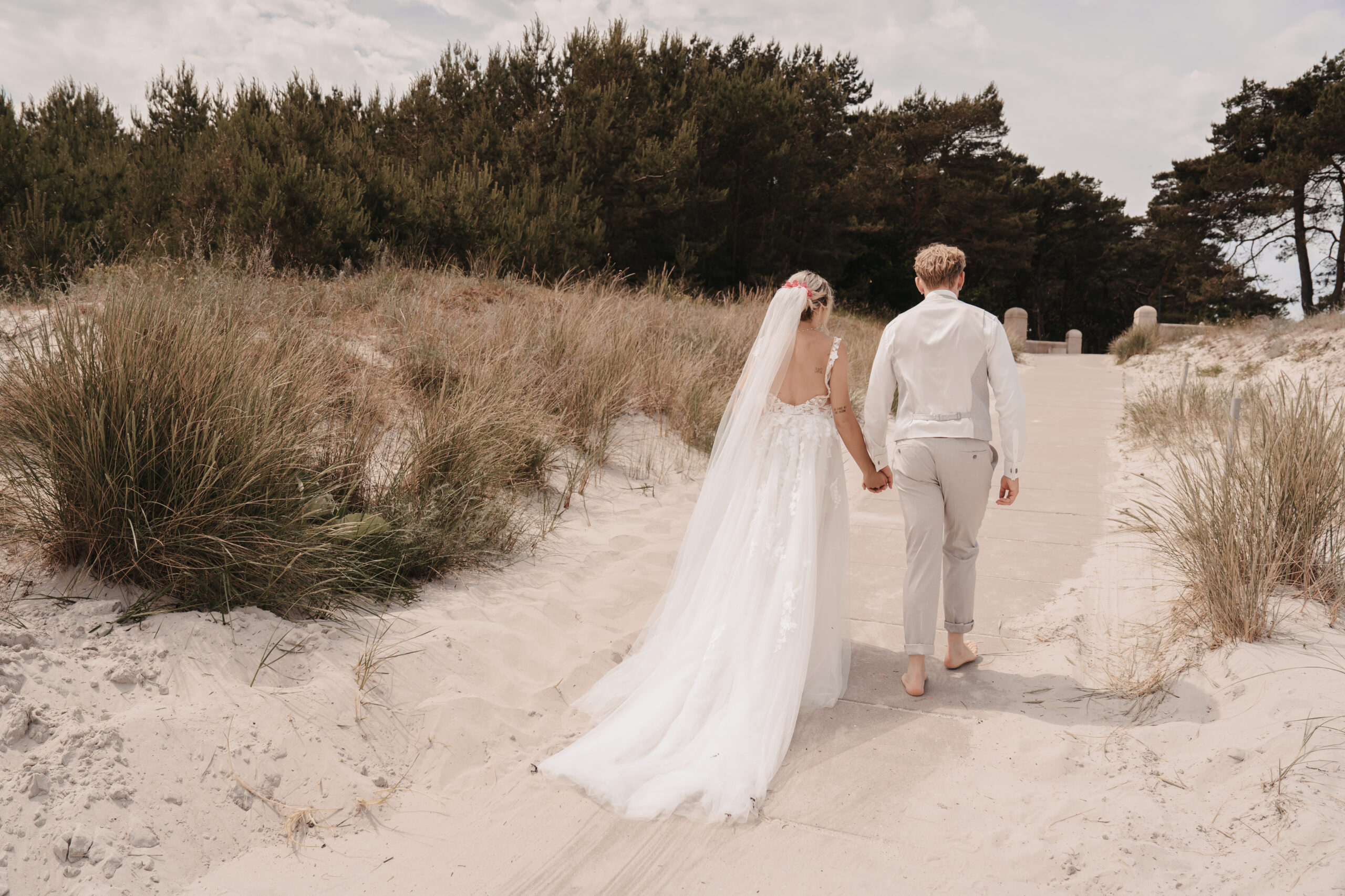 Eine Braut und ein Bräutigam gehen barfuß über einen Sandweg in Richtung Ostseestrand. Sie halten sich an den Händen und sind in Hochzeitskleidung gekleidet.