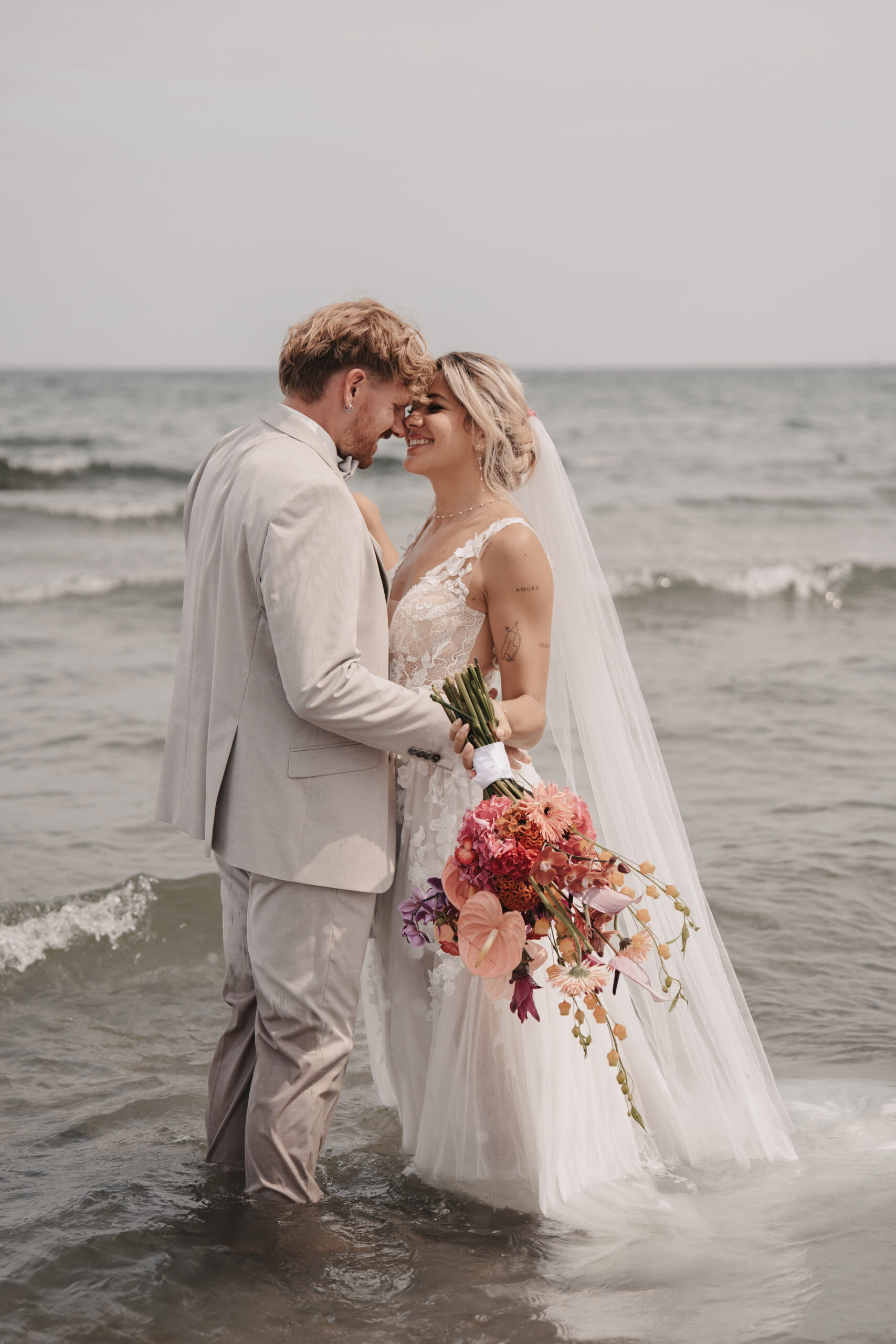 Braut und Bräutigam stehen lächelnd und umarmt im Meer vor Sylt, die Braut hält einen bunten Blumenstrauß - die perfekte Inspiration für jeden Hochzeitsplaner.