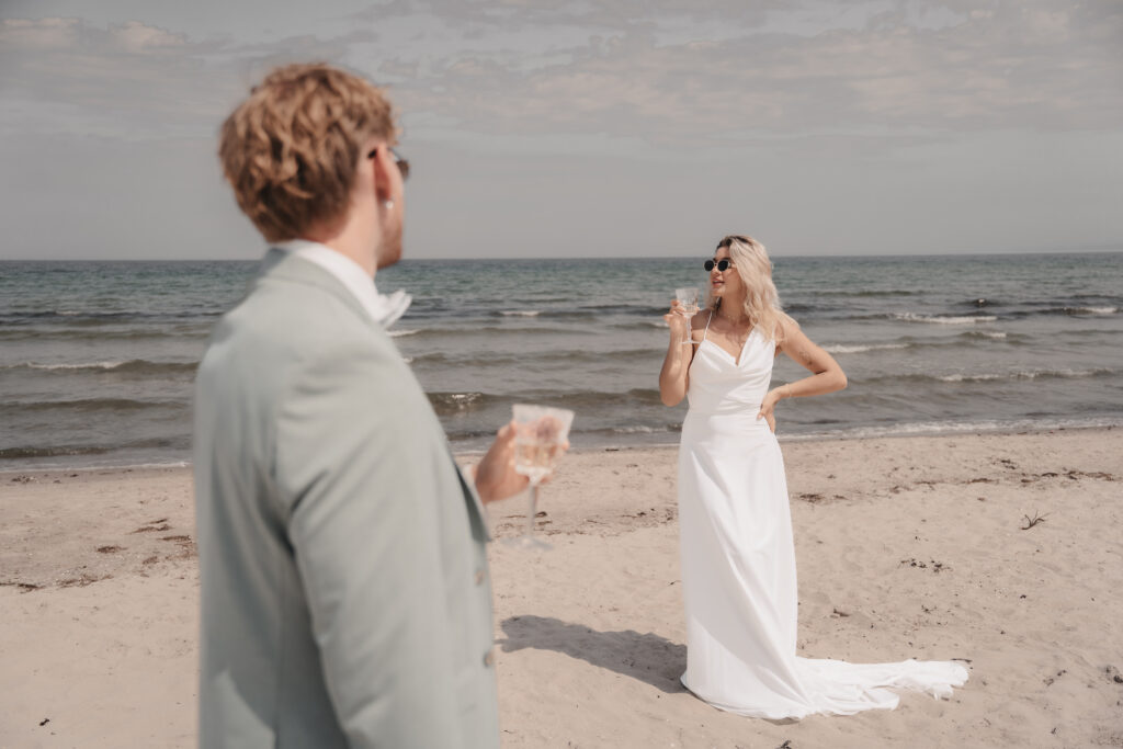 Die Braut im weißen Kleid und der Bräutigam im Anzug trinken Champagner an einem Sandstrand mit dem Meer im Hintergrund, um ihre Hochzeit am Meer zu feiern.
