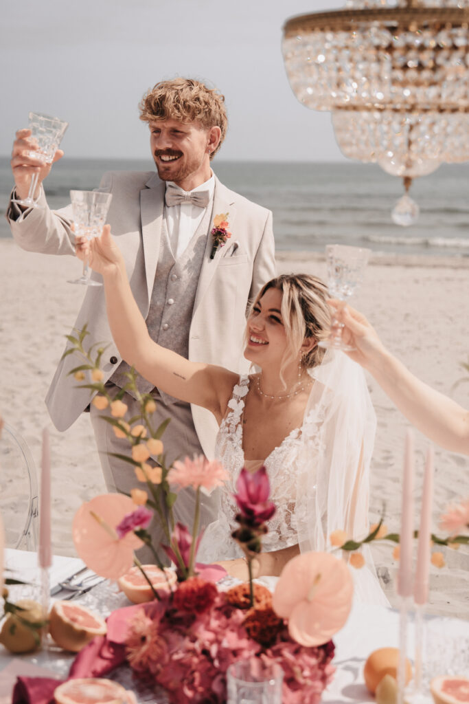 Braut und Bräutigam in Hochzeitskleidung stoßen an einem schön dekorierten Tisch am Strand von Sylt an.