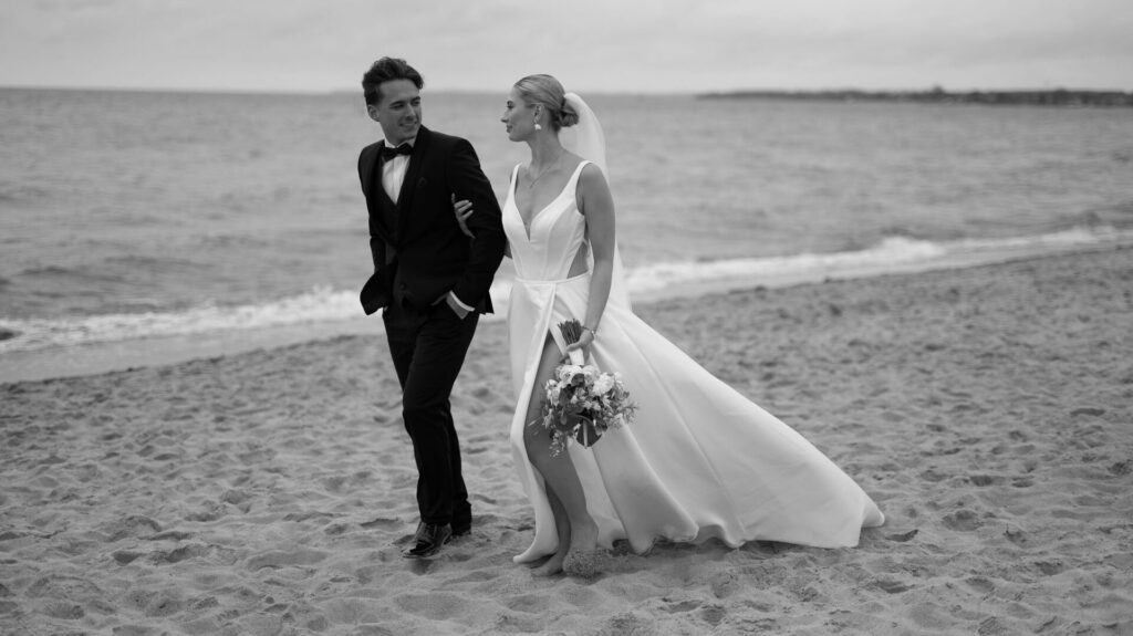 Eine Braut und ein Bräutigam spazieren gemeinsam über einen Sandstrand und lächeln sich am Meer an - perfekt für eine Hochzeit am Meer oder eine romantische Strandhochzeit.