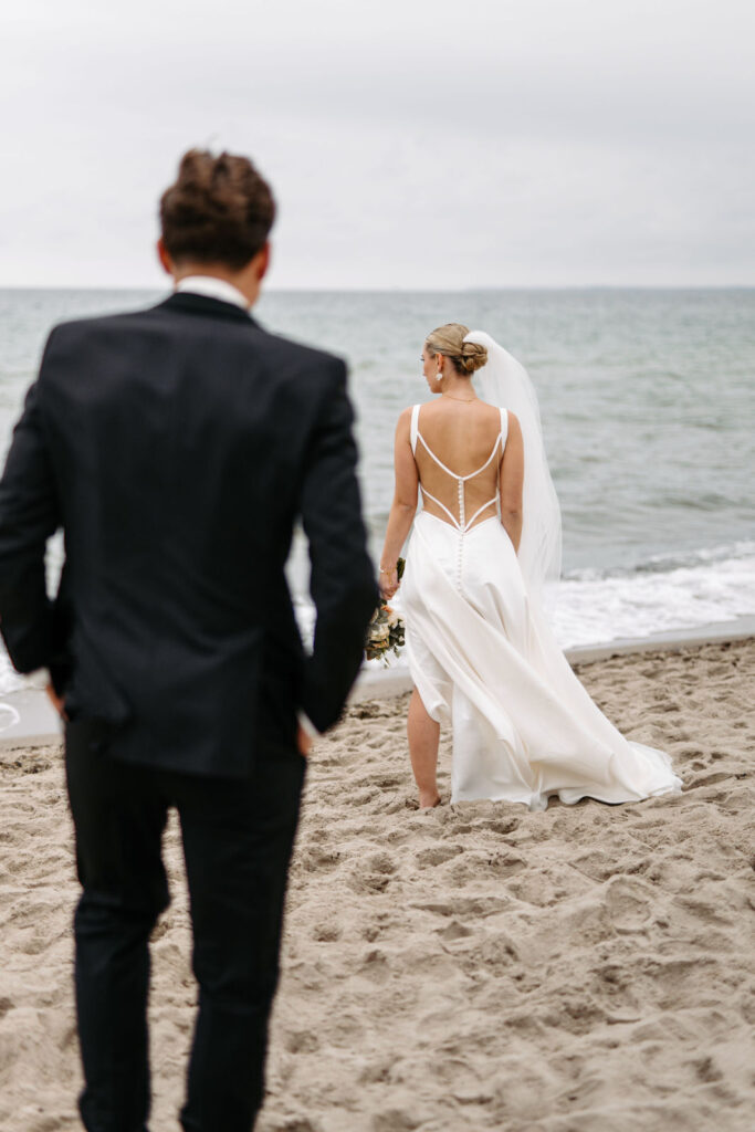 Eine Braut in einem weißen Kleid geht am Strand spazieren, während ein Mann im Anzug zuschaut und den Zauber einer Strandhochzeit mit dem Meer im Hintergrund festhält.