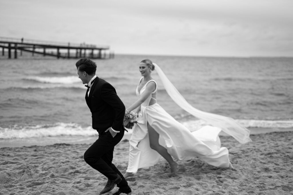 Eine Braut jagt spielerisch einen Bräutigam an einem Sandstrand in der Nähe des Meeres, beide lächelnd und formell gekleidet - der perfekte Moment für eine Strandhochzeit.