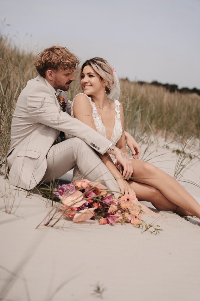 Braut und Bräutigam sitzen eng beieinander auf einer Sanddüne und lächeln sich mit einem bunten Blumenstrauß an - ein perfekter Moment für eine Strandhochzeit oder Hochzeit am Meer.