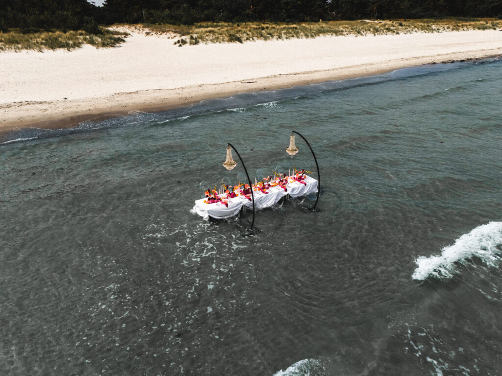 Ein langer, geschmückter Tisch ist im flachen Meerwasser in der Nähe eines Sandstrandes aufgestellt, an dem Menschen speisen - perfekt für eine unvergessliche Hochzeit am Meer.