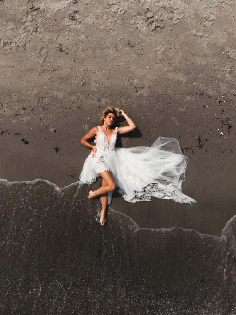 Eine Frau im weißen Hochzeitskleid liegt am Sandstrand in der Nähe der Wasserkante und fängt den Zauber einer Hochzeit am Meer ein.