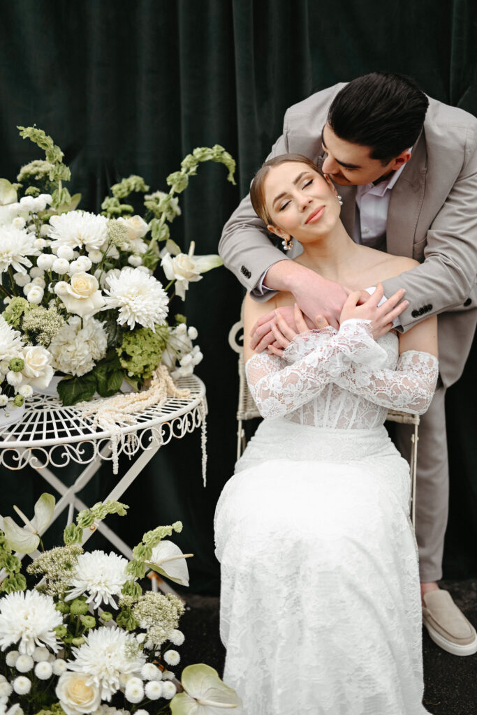 Ein Bräutigam umarmt und küsst seine Braut, die gelassen in einem weißen Spitzenkleid neben Blumenarrangements sitzt - ein unvergesslicher Moment am Ufer der Ostsee, perfekt inszeniert von einem erfahrenen Hochzeitsplaner.