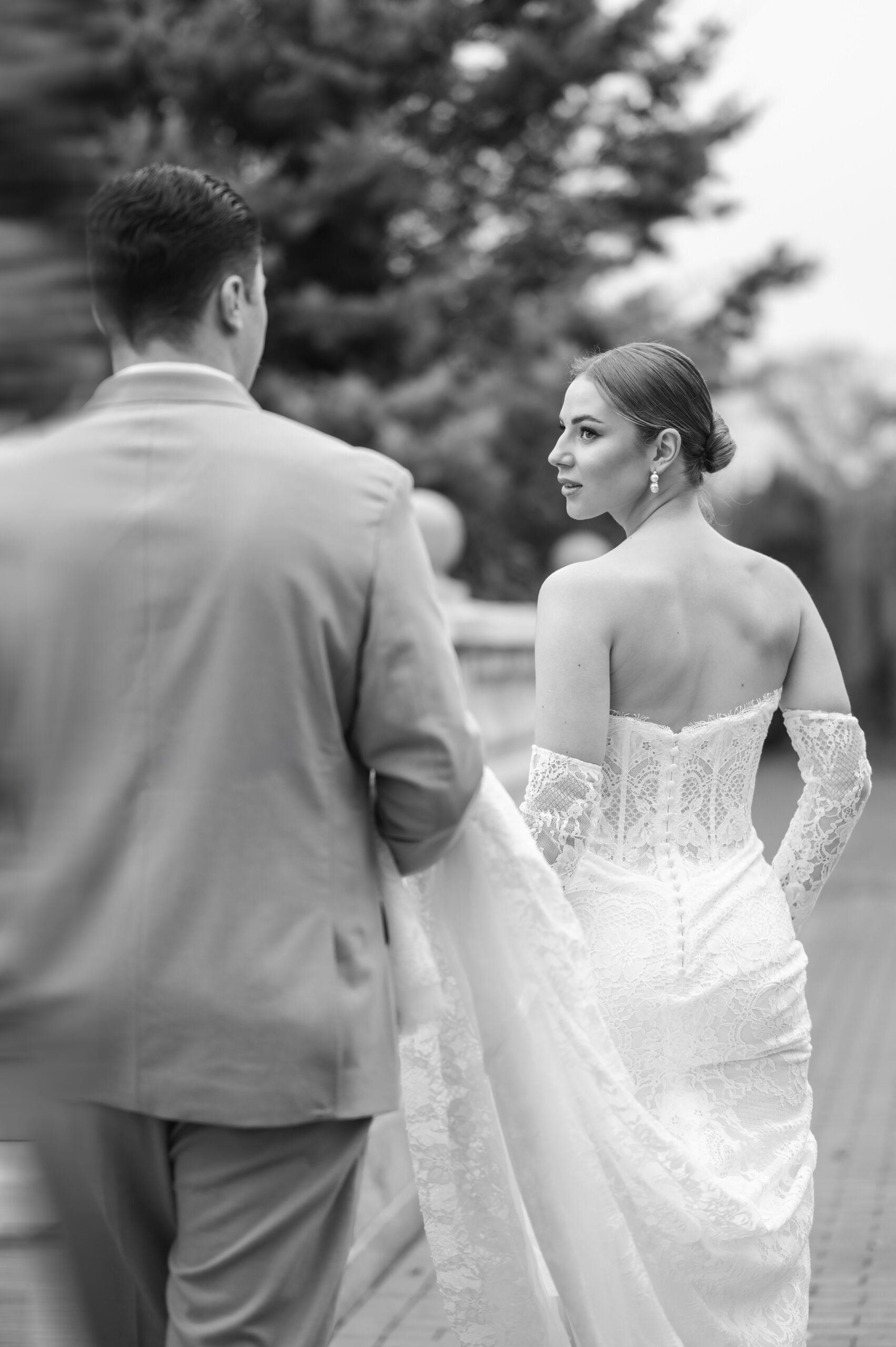 Eine Braut im Spitzenkleid blickt auf ihren Bräutigam, der ihre Schleppe hält, während sie im Freien an der malerischen Küste Rügens spazieren gehen - perfekt für alle Hochzeitsplaner, die Inspiration an der Ostsee suchen.