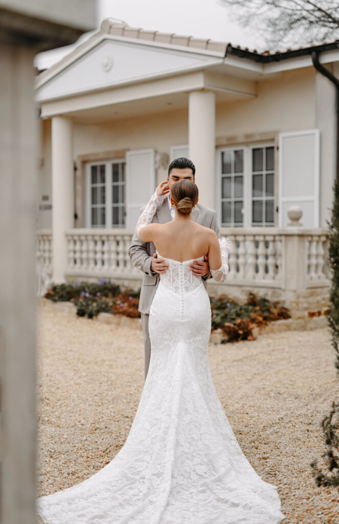 Braut und Bräutigam umarmen sich vor einem weißen Haus auf Rügen; die Braut trägt ein Spitzenkleid mit langer Schleppe, perfekt für die von einem erfahrenen Hochzeitsplaner kuratierten Erinnerungen.