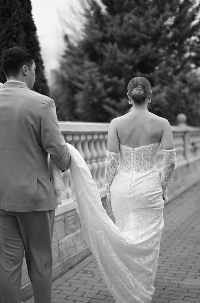 Eine Braut im Spitzenkleid schreitet mit ihrem Bräutigam, der ihre Schleppe hält, einen gemauerten Weg im Freien entlang - die perfekte Inspiration für Paare, die einen Hochzeitsplaner auf Sylt oder eine elegante Feier an der Ostsee planen.