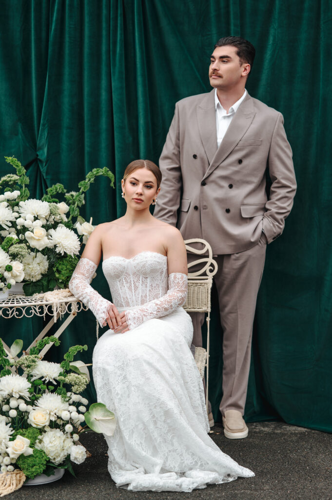 Eine Braut im Spitzenkleid sitzt zwischen Blumen, der Bräutigam im hellbraunen Anzug steht neben ihr vor einem grünen Vorhang - die perfekte Inspiration für Paare, die von einer Ostsee-Hochzeit auf Rügen träumen, begleitet von einem erfahrenen Hochzeitsplaner.