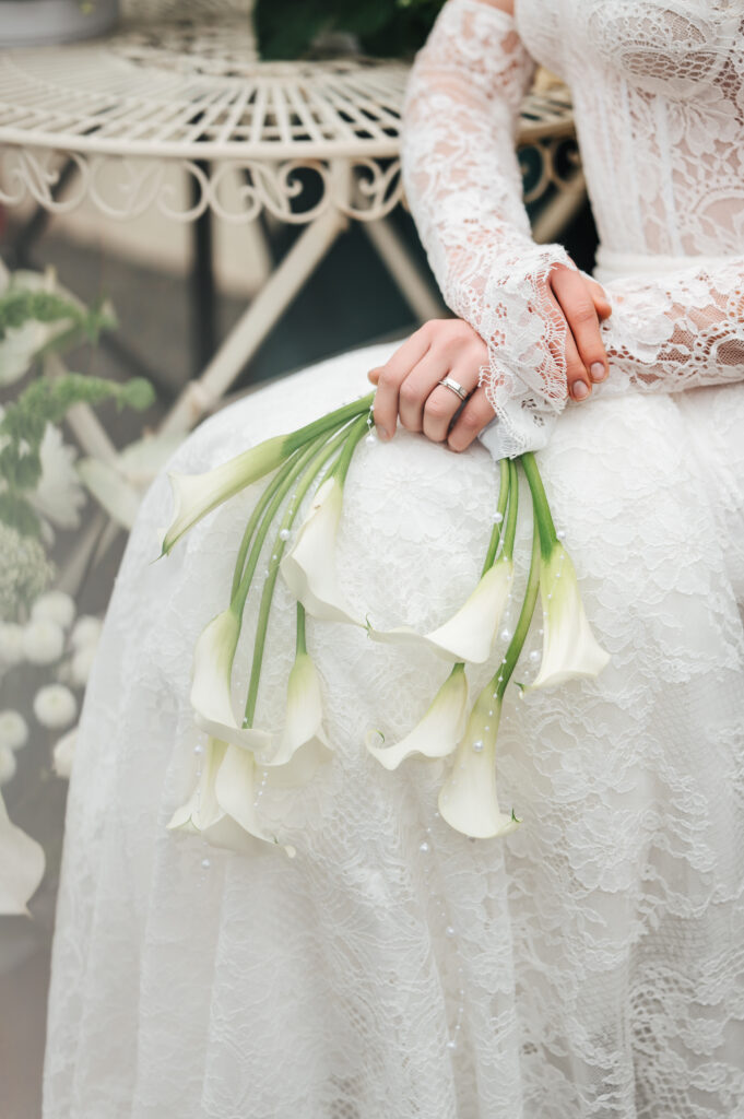 Braut im Spitzenhochzeitskleid hält weiße Calla-Lilien auf Sylt und zeigt ihren Verlobungsring.