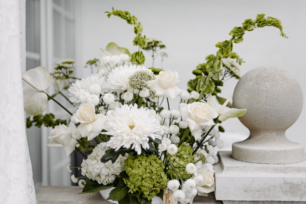 Ein weißes und grünes Blumenarrangement auf einem Steinsims neben einem runden Baluster, das an die frische Eleganz von Sylt erinnert - die perfekte Inspiration für jeden Hochzeitsplaner, der den Charme der Ostsee sucht.