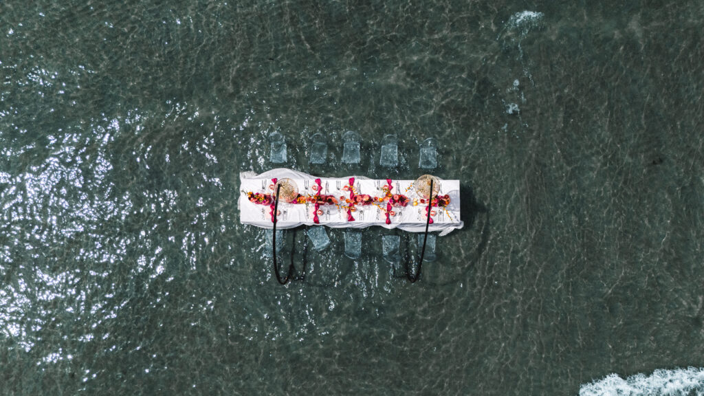Luftaufnahme eines dekorierten Esstisches mit Stühlen, der im flachen Meerwasser aufgestellt ist, perfekt für eine Strandhochzeit oder für alle, die von einer Hochzeit am Meer träumen.