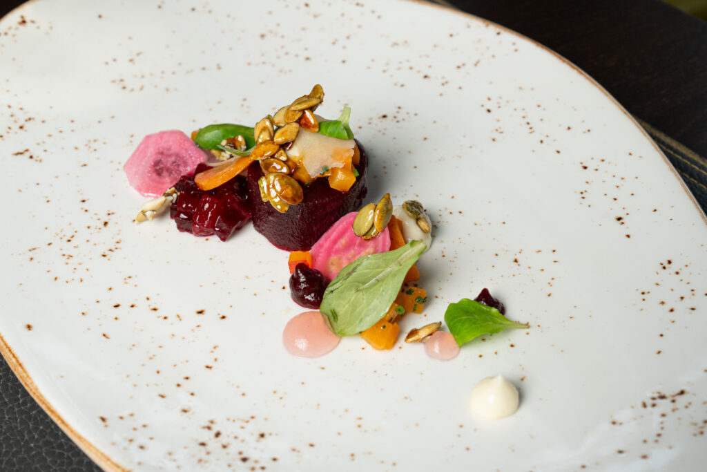 Ein Gourmet-Gericht mit buntem Gemüse, Samen und Soße, das kunstvoll auf einem weiß gesprenkelten Teller angerichtet ist.