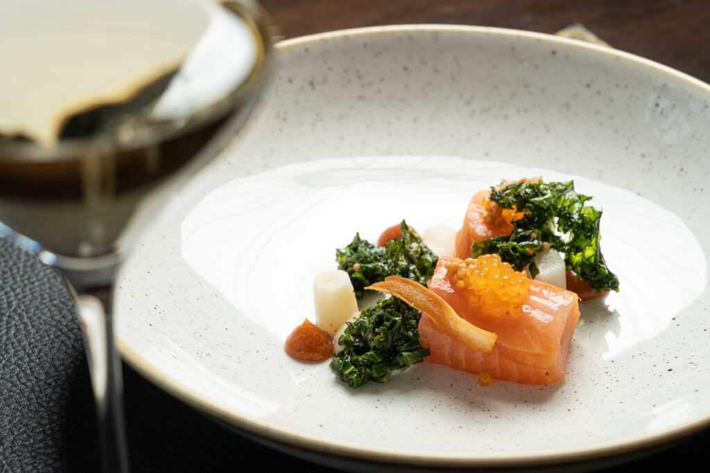Ein Gourmet-Gericht mit Lachs, Grünzeug, weißen Würfeln und orangefarbenem Rogen auf einem weißen Teller; ein Getränk steht unscharf davor.