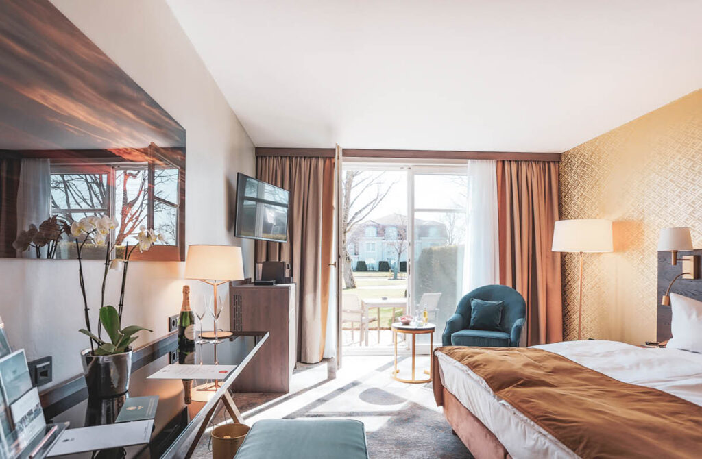 Helles Hotelzimmer mit Bett, Schreibtisch, TV, Sessel und Glastüren zum sonnigen Innenhof und Garten.