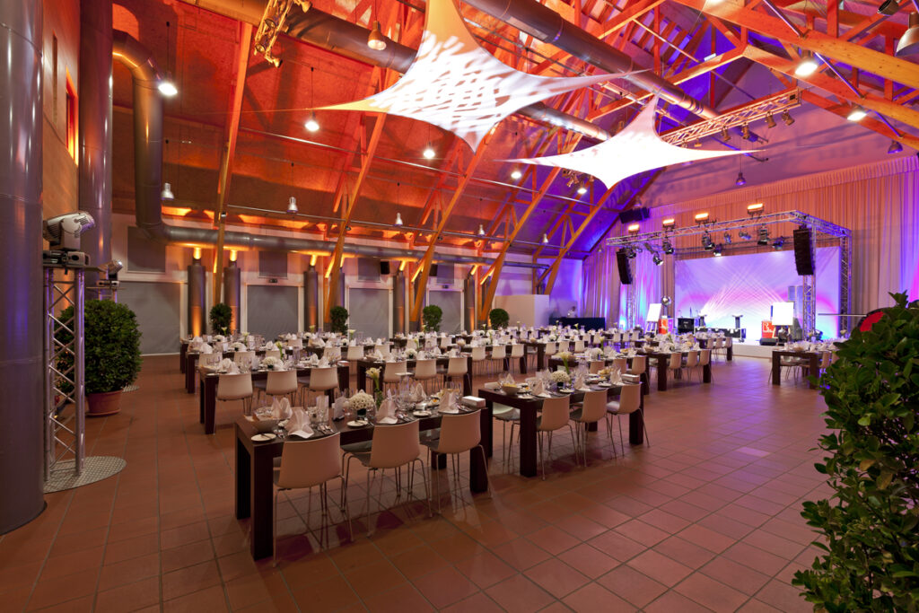 Großer, eleganter Veranstaltungssaal mit dekorierten Tischen, Bühne, dramatischer Beleuchtung und moderner Architekturdecke.