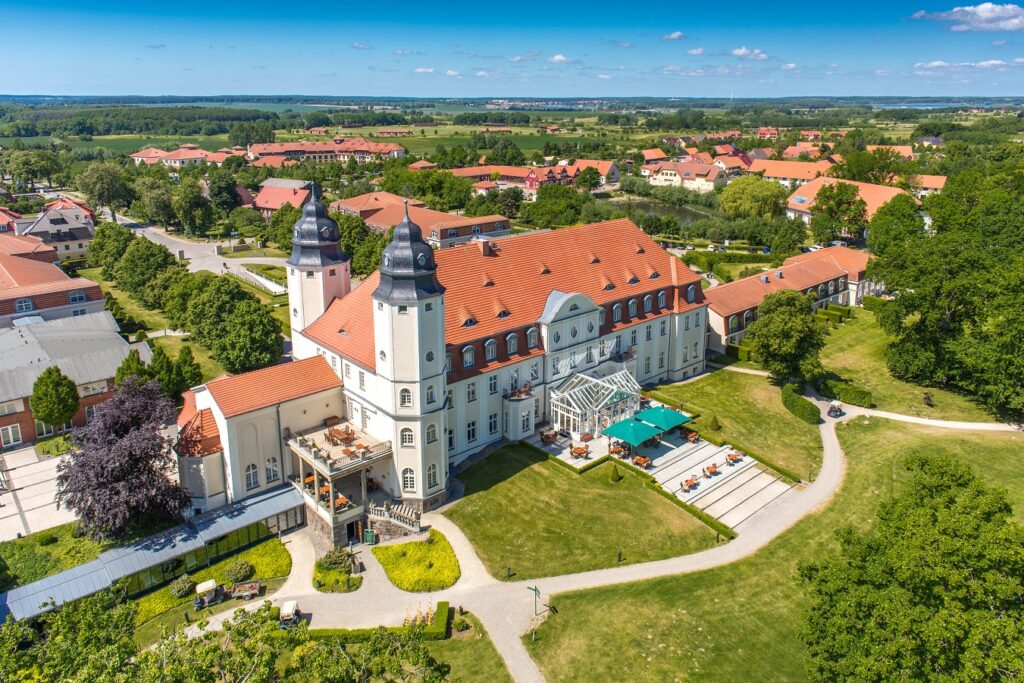 Luftaufnahme von Schloss Fleesensee, einem großen, eleganten Gebäude mit roten Dächern, Türmen und umliegenden grünen Gärten an einem sonnigen Tag.