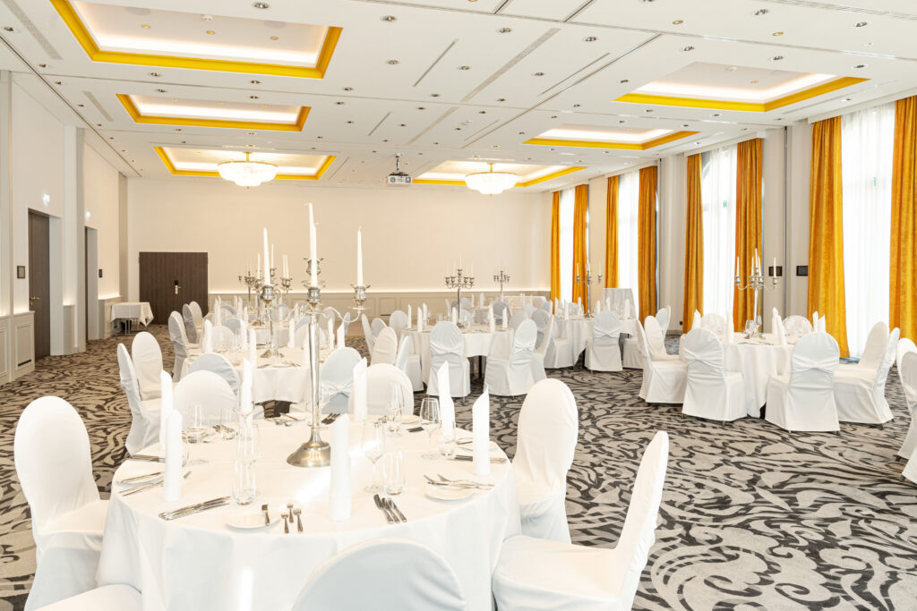 Eleganter Bankettsaal mit runden Tischen, weißen Stühlen und hohen Kerzenständern, beleuchtet von großen Fenstern und Kronleuchtern.