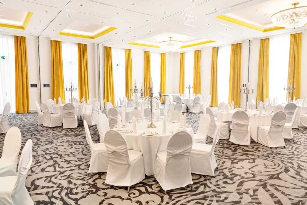 Ein heller Bankettsaal mit runden Tischen, weißen Stühlen, gelben Vorhängen und eleganten Kronleuchtern.