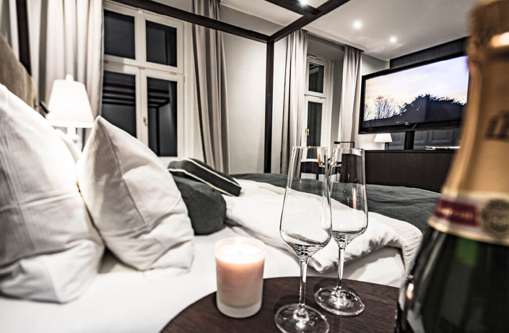 Elegantes Hotelzimmer mit einem gemütlichen Bett, einer Kerze, Champagner, zwei Gläsern und einem Fernseher im Hintergrund.