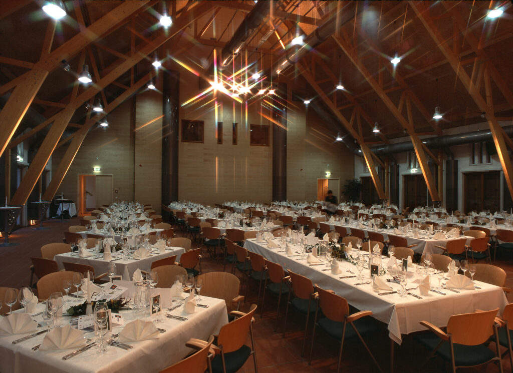 Großer Bankettsaal mit Holzbalken, gedeckte Tische mit weißen Tüchern, Gläsern und Servietten, bereit für eine Veranstaltung.