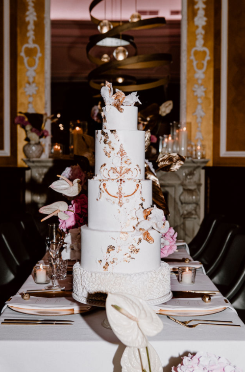 Große, verschnörkelte weiße Hochzeitstorte mit goldenen Blumendetails auf einem eleganten, dekorierten Esstisch.