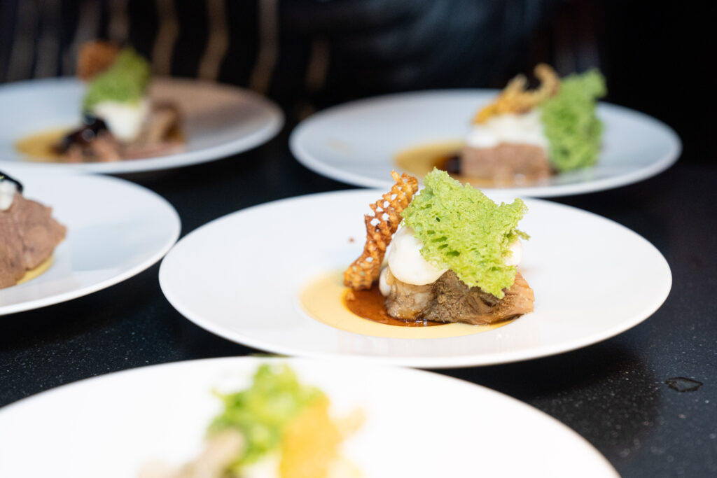 Ein plattiertes Gourmetgericht mit Fleisch, grüner Beilage, Soße und einem knusprigen Element, serviert auf einem weißen Teller.