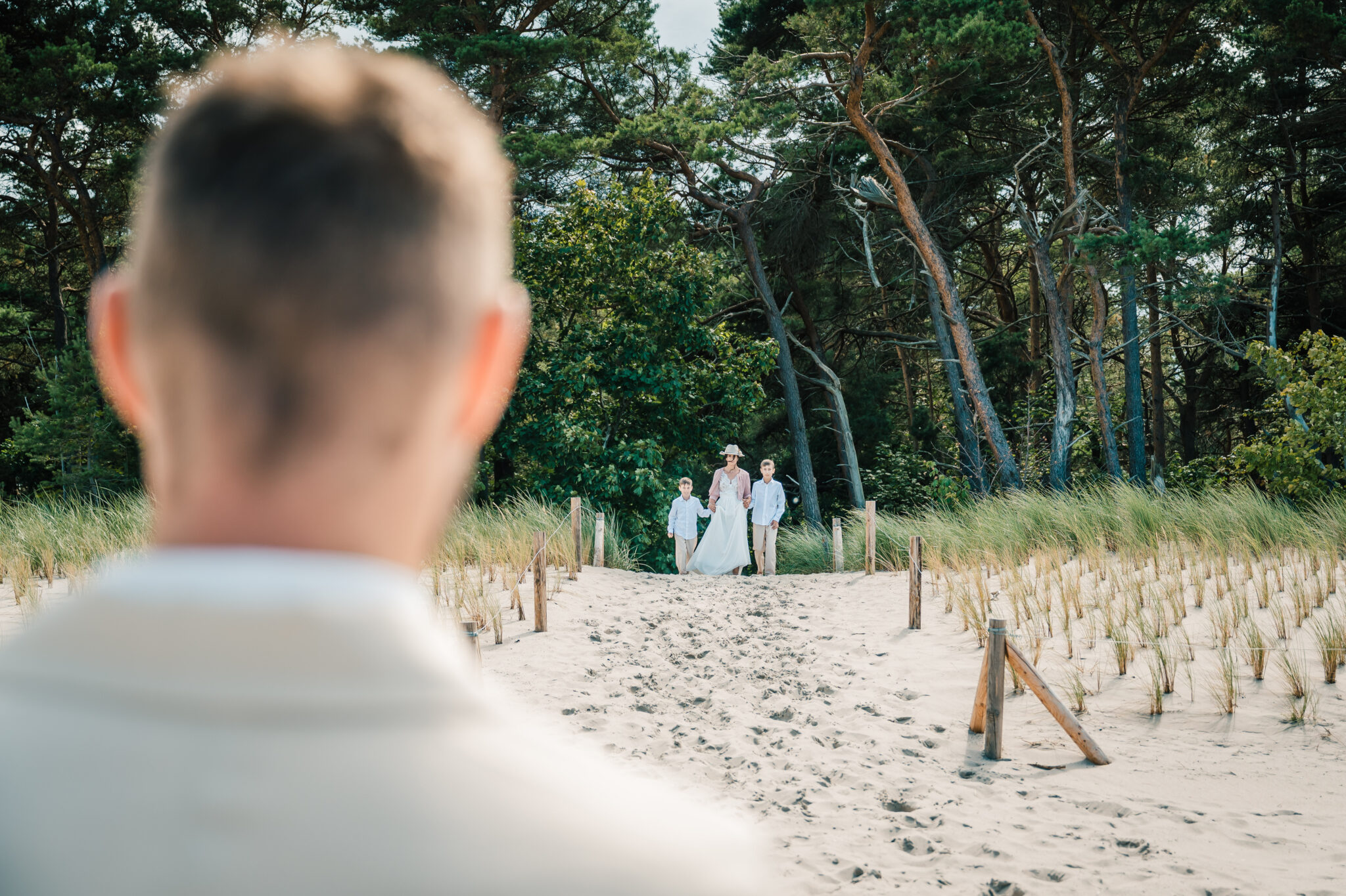 Eine Person in weißer Kleidung beobachtet eine Braut und ihre Begleiter, die bei einer Hochzeit im Freien einen sandigen, grasbewachsenen Weg entlanggehen.